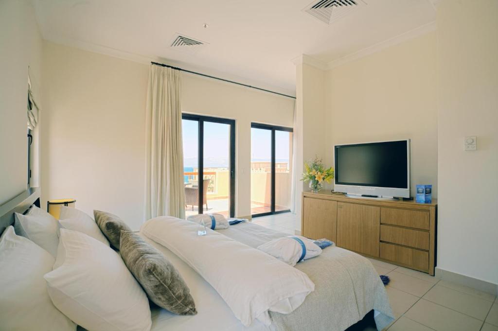 Mūsdienīgs guļamistabas interjers ar plašu logu un televizoru. Gultā ir ērti spilveni, bet blakus atrodas koka kumode un ziedu kompozīcija.