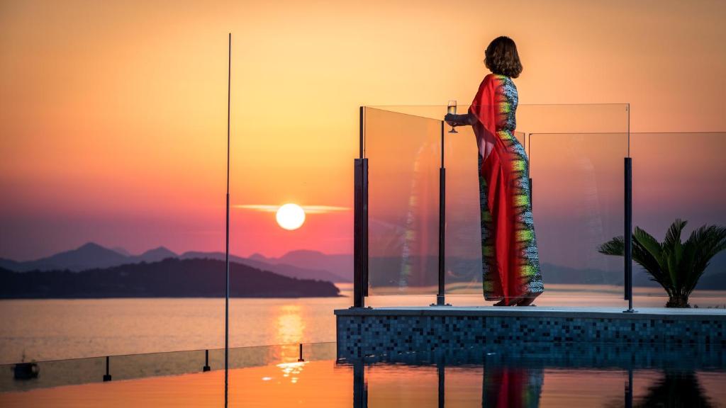 Sieviete krāsainā kleitā, stāvot uz terases pie baseina, vēro saulrietu. Fons ir krāšņs jūras skats un kalni, kas apgaismoti ar saulrieta krāsām.