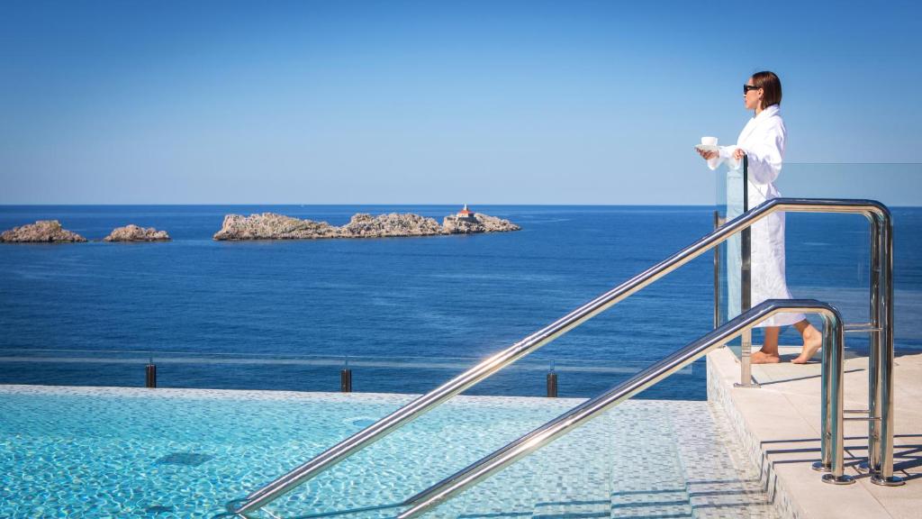 Sieviete peldkostīmā stāv pie peldbaseina, baudot skatu uz jūru un akmeņainām salām. Viņa tur rokās tasi, izskatoties relaksēta un apmierināta.