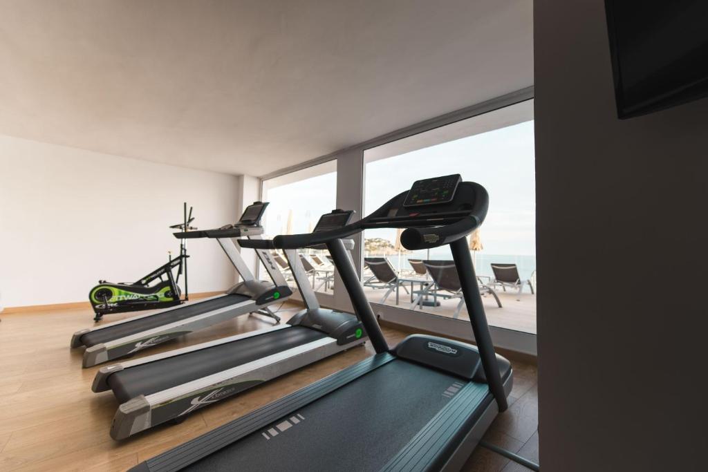Mūsdienīgs sporta zāles interjers ar trīs skrejceliņiem un velosipēdu, kas atrodas blakus plašam logam ar skatu uz āra baseinu un sauļošanās krēsliem.