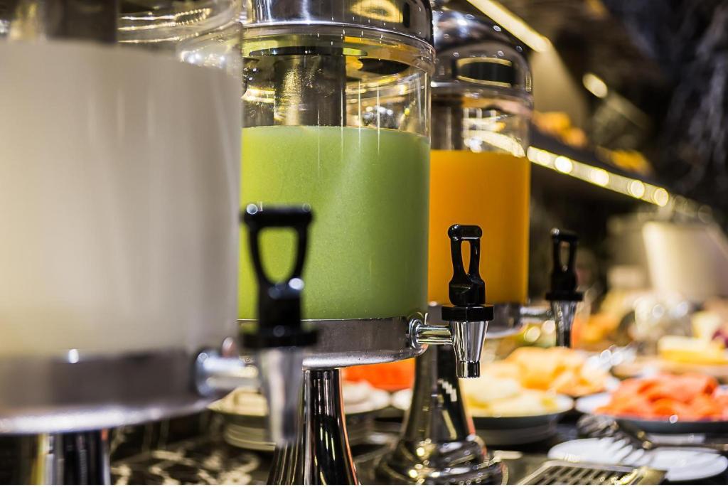 Svaigu dzērienu dispensers ar dažādām krāsām, tostarp zaļš un oranžs, novietoti uz galda, blakus dažādiem ēdieniem.