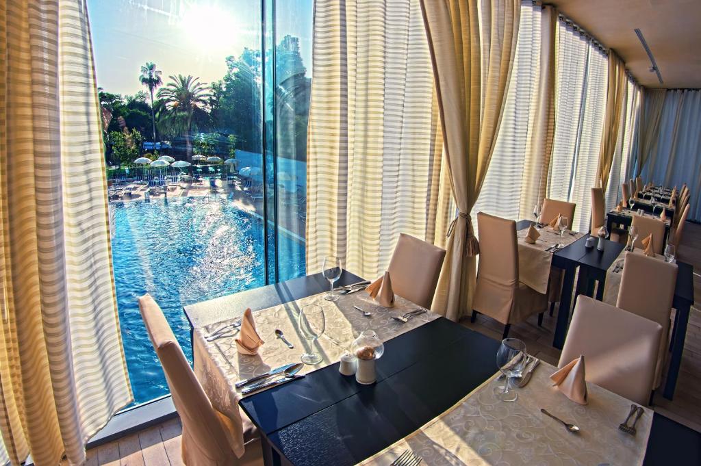 Labi iekārtota restorāna interjers ar skatu uz baseinu un sauli, kas spīd caur logiem. Elegantās galda uzkopes un gaišas aizkaru krāsas veido patīkamu atmosfēru.