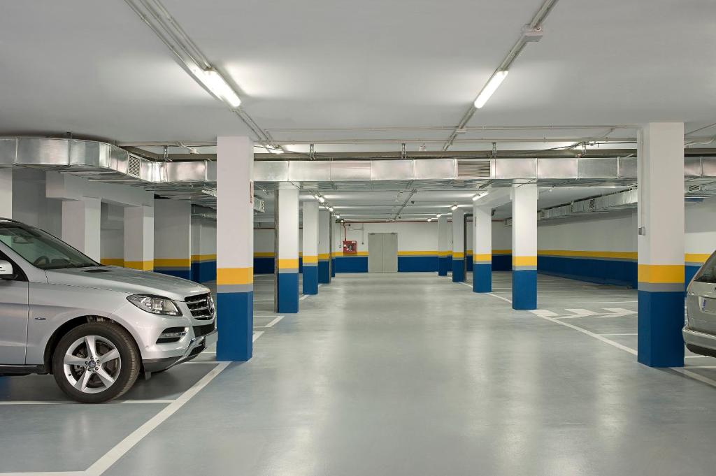 Lielas, tukšas autostāvvieta ar pelēku automašīnu un krāsotām kolonām zili dzeltenos toņos. Apgaismojums ir spilgts, radot tīru un modernu atmosfēru.
