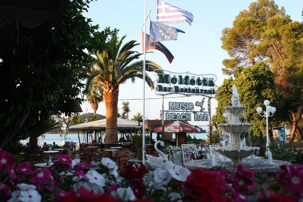 Molfetta bārs un restorāns pie pludmales, apkārt palmām un ziediem. Attēlā redzama strūklaka un karogi, kas plīvo virs ēkas.