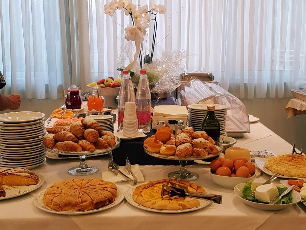 Bagātīgs brokastu galds ar dažādiem konditorejas izstrādājumiem, augļiem un dzērieniem. Uz galda redzami kruasāni, kūkas, vārītas olas un salāti.