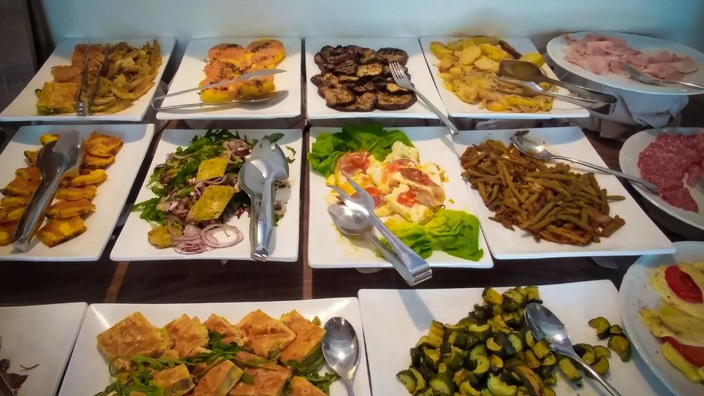 Dažādu ēdienu bufete ar salātiem, dārzeņiem un gaļu uz baltām šķīvju plāksnēm. Izvēle ietver ceptus dārzeņus, salātus un gaļas delikateses.