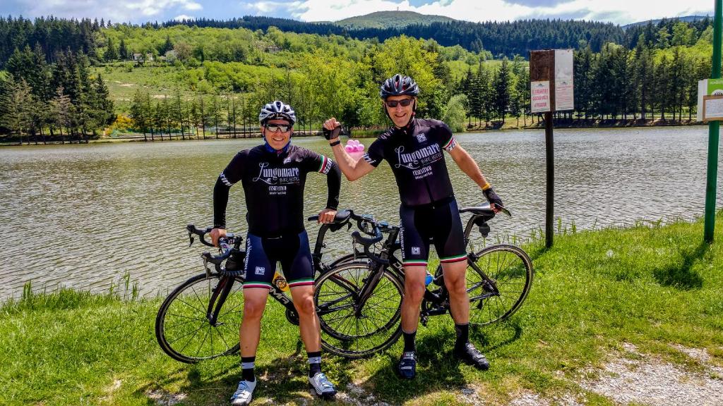 Divi riteņbraucēji pozē pie ezera, tērpti sporta apģērbu un ķiverēm, ar velosipēdiem blakus. Fona ainava ietver mežainus kalnus un skaidru debesi.