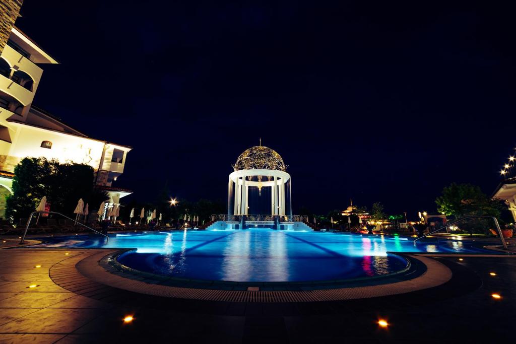 Vakara skats uz elegantu baseinu ar apgaismotu paviljonu un apkārtējiem kokiem, radot mierīgu un relaksējošu atmosfēru.