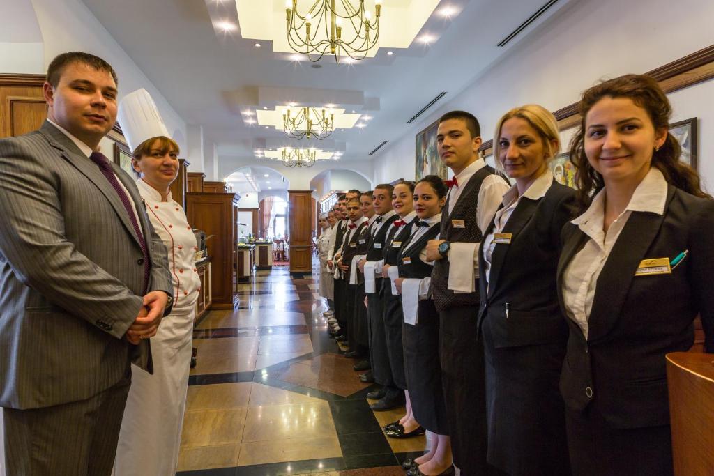 Restorāna darbinieku grupas attēls, kas stāv rindā eleganti noformētā telpā, uzsverot profesionālo apkalpošanu. Fons ietver gaumīgi dekorētas telpas elementus un apgaismojumu.