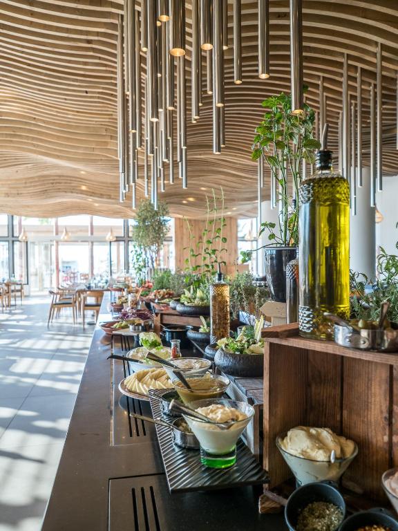 Mūsdienīgs restorāns ar koka interjeru un elegantiem gaismas elementiem, kurā redzama plaša ēdienu izvēle, tostarp salāti un mērces. Gaisīgā atmosfērā ir daudz augiem un dabīgām dekorācijām.