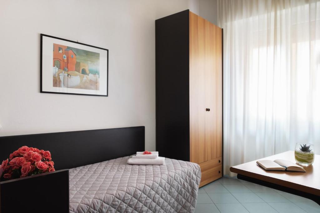 Mūsdienīga guļamistaba ar gultu, ziedu kompozīciju un modernu mēbeļu dizainu, kas rada mājīgu atmosfēru. Sienā ir piekārta mākslas gleznas kopija, un telpā ieplūst dabiska gaisma caur baltām aizkarām.