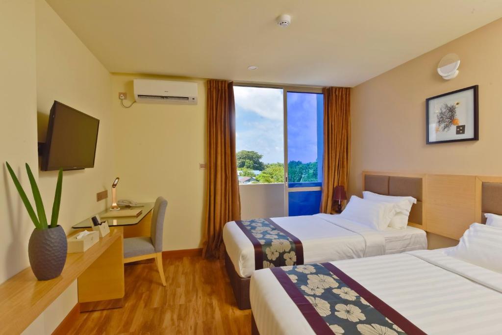 Mūsdienīga viesnīcas istaba ar divām gultām, darba galdu un televizoru, kurā plūst dabiska gaisma caur lielajiem logiem. Loga priekšā redzama ainava ar zaļiem kokiem.