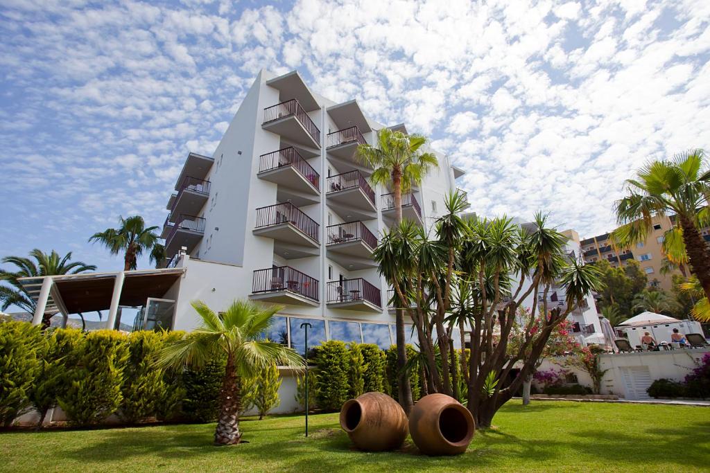 Moderns viesnīcas ēka ar balkoniem, ieskauta tropiskā dārzā ar palmām un dekoratīviem podiem. Skaista debess ar mākoņiem papildina ainavu.