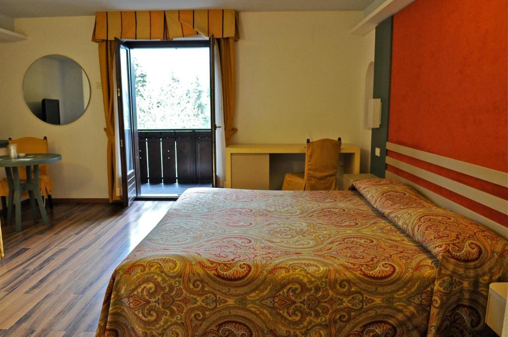 Gaiša viesnīcas numura iekārtojums ar divguļamo gultu, koka grīdu un balkonu. Logā redzams zaļš skats, un istabā ir neliels galds ar krēsliem.