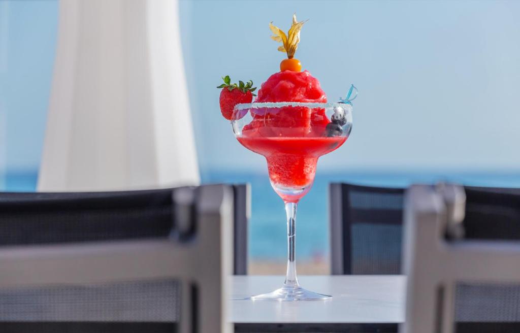 Svaigs un krāsains kokteilis ar zemeņu un augļu sasalumu, pasniegts caurspīdīgā glāzē, ar jūras ainavu fonā.