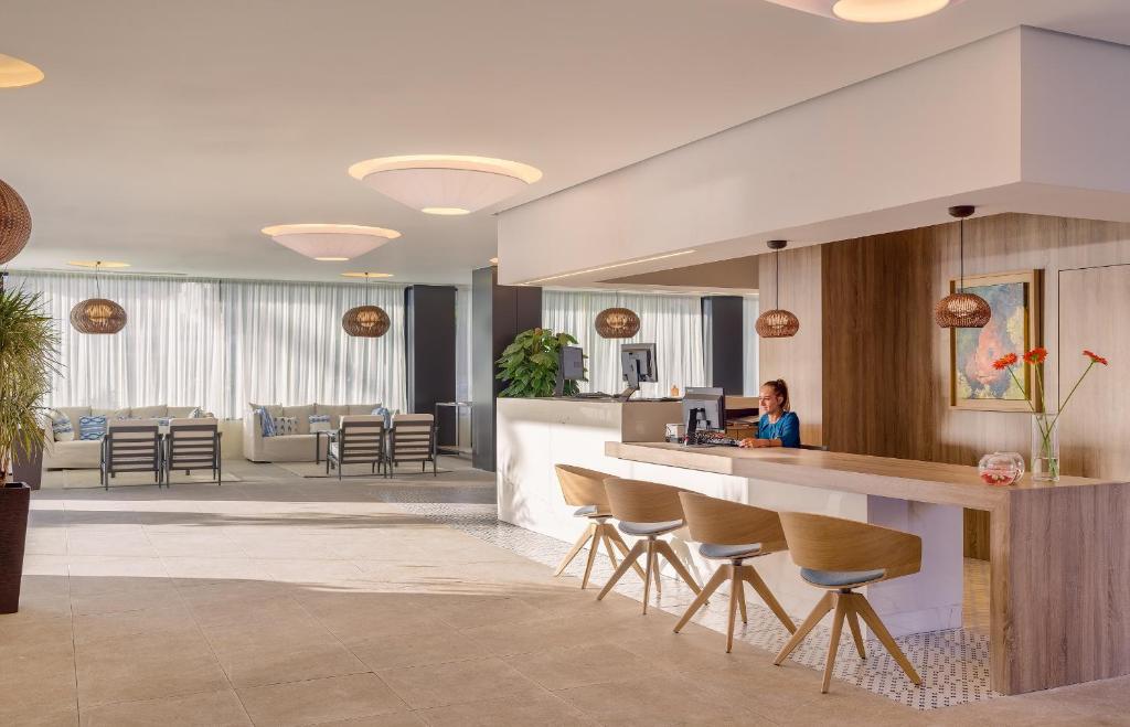Mūsdienīgs viesnīcas reģistratūras interjers ar apgaismojumu un komfortablām mēbelēm, kurā redzama darbiniece pie galda.