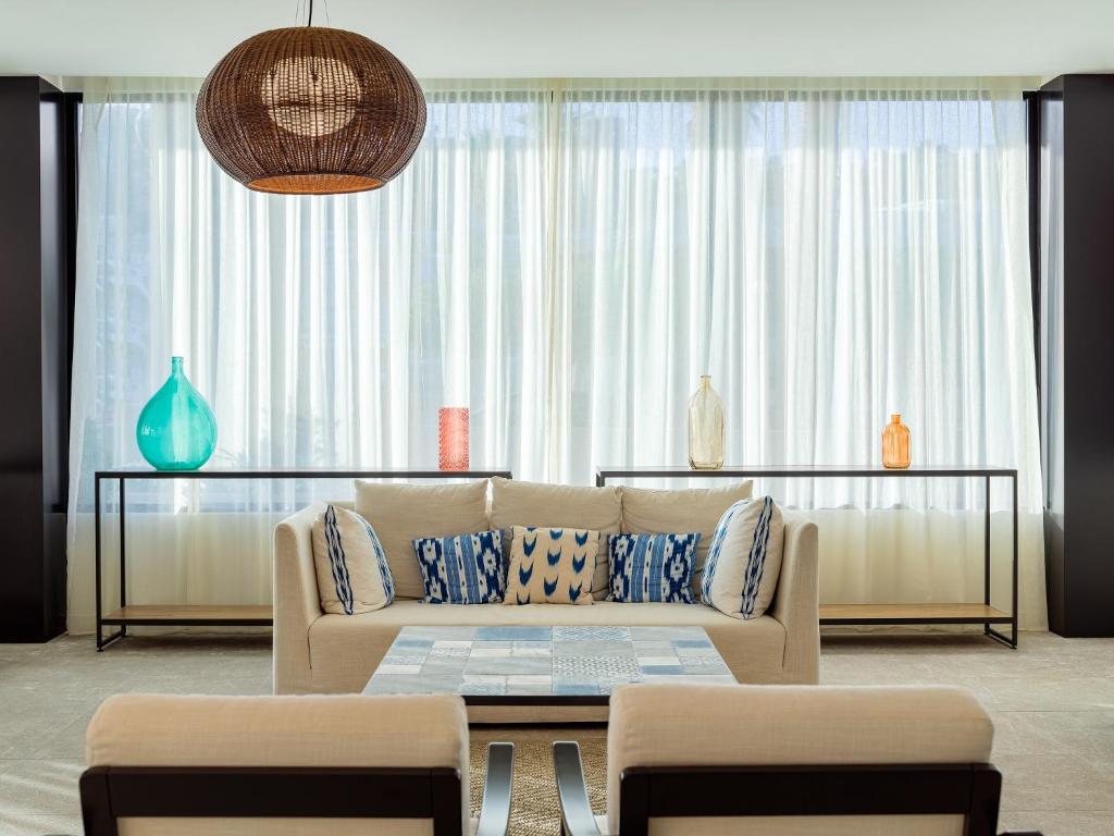 Mūsdienīgs viesistabas interjers ar gaisīgu apgaismojumu, baltām aizkariem un krāsainām stikla vīzēm uz plauktiem. Komfortablas dīvāna un krēslu kombinācijas ar rakstainiem spilveniem.