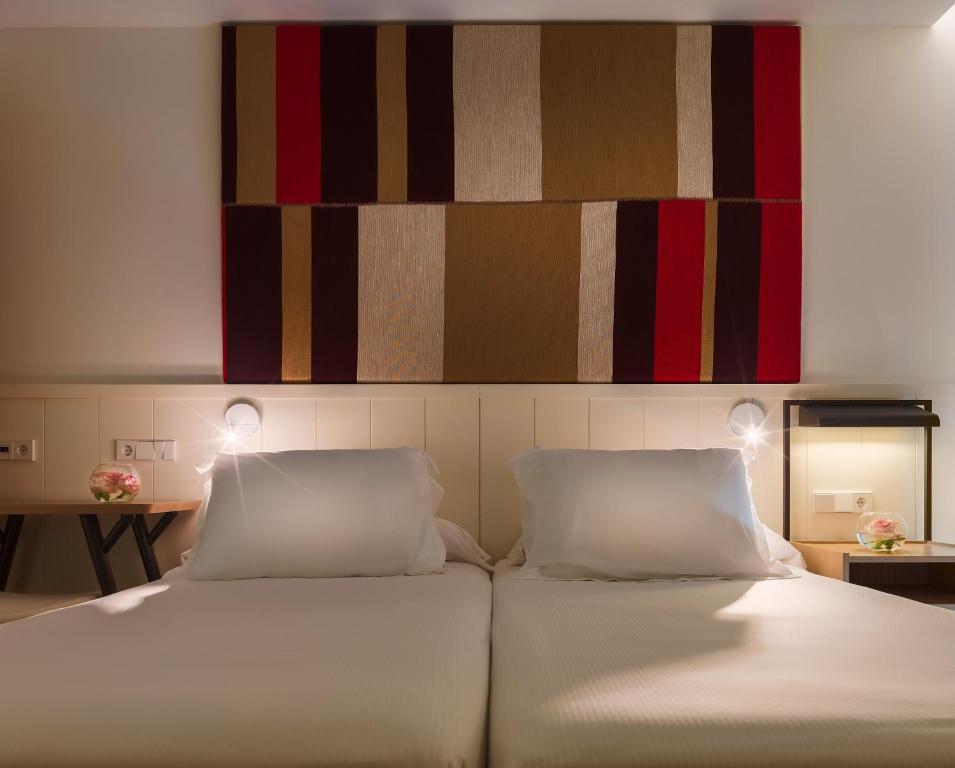 Mūsdienīgs guļamistabas interjers ar diviem gultām, pie kura sienas ir krāsains audums. Uz naktsgaldiņa atrodas gaismas avots un ziedu kompozīcija.