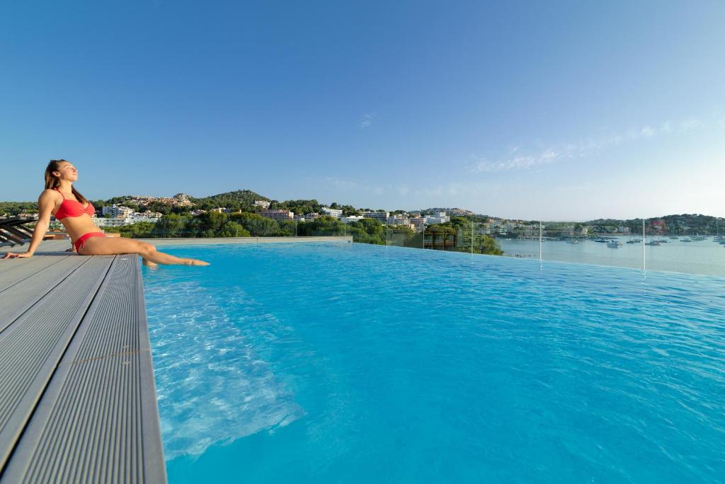 Sieviete sarkanos peldkostīmos relaksējas pie bezgalības baseina, baudot sauli un skatu uz kalniem un jūru.