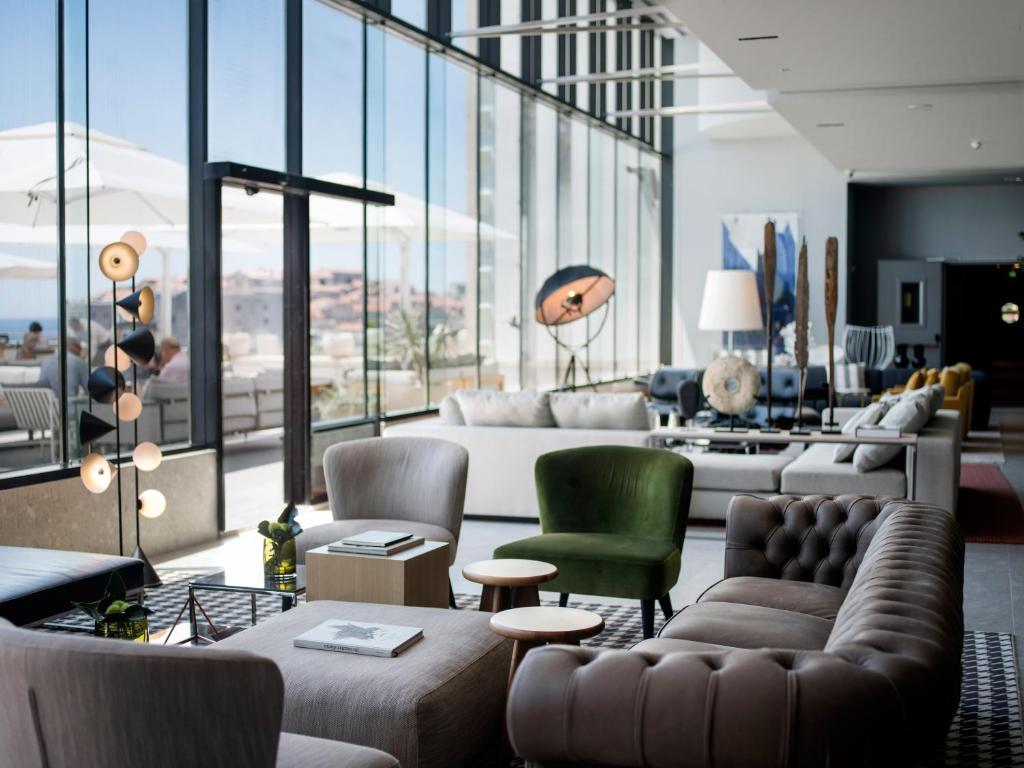 Mūsdienīgs viesnīcas viesistabas interjers ar lieliem logiem, kas sniedz skatu uz ārpusi. Telpā ir dažādas mēbeles, tostarp ērti krēsli un dīvāni, kā arī dekoratīvi elementi.