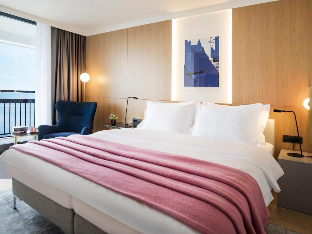 Mūsdienīgs viesnīcas numurs ar plašu gultu, rozā segu un skatu uz jūru. Interjers apvieno koka un mīksto audumu elementus, radot relaksējošu atmosfēru.