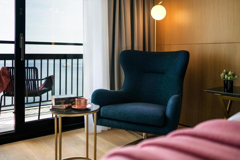 Mūsdienīgs viesnīcas numurs ar skatu uz jūru, kurā ir ērta zila krēsla un stilīga galda kompozīcija ar kafijas krūzi un grāmatām.
