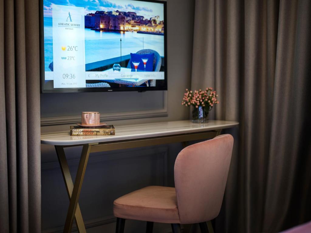 Moderns viesnīcas numura interjers ar elegantu galdu, rozā krāsas krēslu un televizoru, kas rāda laika prognozi. Uz galda atrodas grāmatas un pušķis ar ziediem.