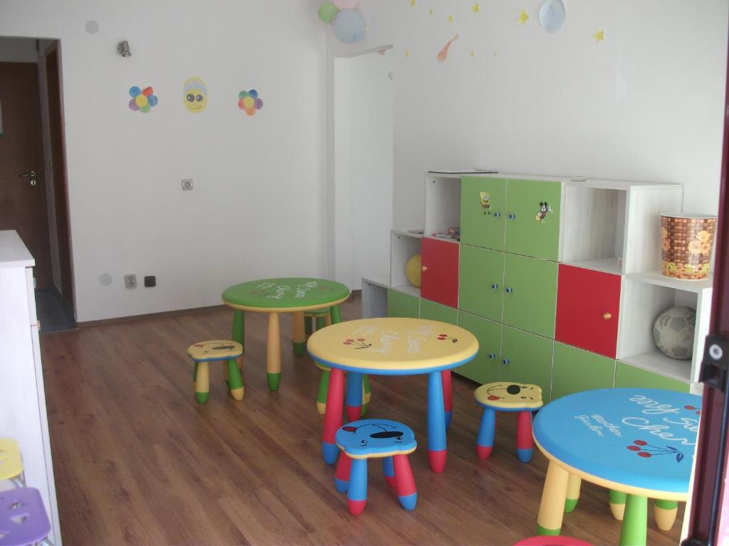 Krāsains bērnu istabas interjers ar vairākām mazām galdiem un krēsliem, kas dekorēti ar jautrām ilustrācijām. Telpā ir arī skapītis ar krāsainām durvīm un rotaļlietām.
