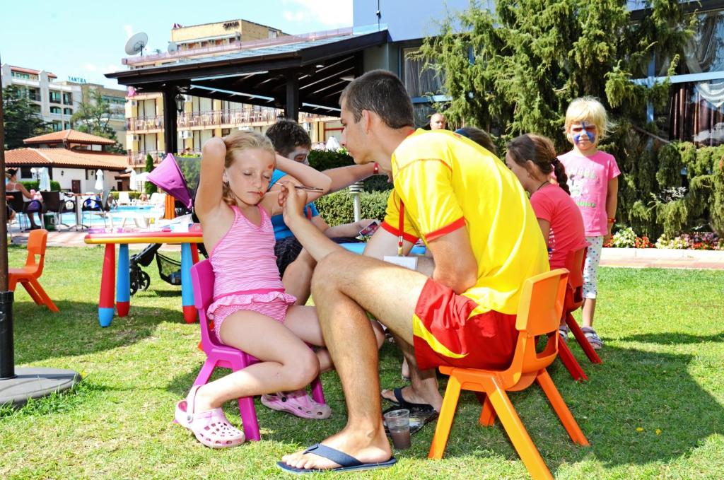 Bērni spēlējas dārzā, kur viens no pieaugušajiem, tērpies peldētāja tērpā, piedalās aktivitātēs un sniedz palīdzību. Apkārt redzami krāsaini krēsli un peldbaseins fonā.