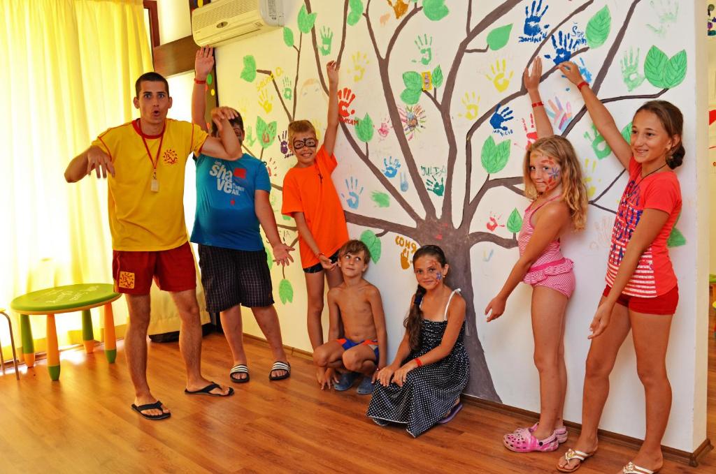 Bērni un vadītājs pozē pie sienas, kurā ir zīmēts koks ar roku nospiedumiem un lapām. Telpa ir gaiša un draudzīga, radot radošu atmosfēru.