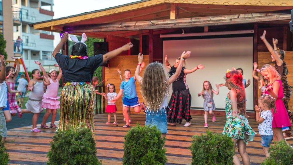Bērni izklaidējas un dejo uz skatuves, kamēr pieaugušie piedalās svinībās, radot priecīgu un jautru atmosfēru.