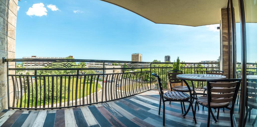 Balkons ar skatu uz zaļo dārzu un pilsētas ainavu, ap kuru novietoti galdi un krēsli. Saulains laiks un zils debesis rada patīkamu atmosfēru.