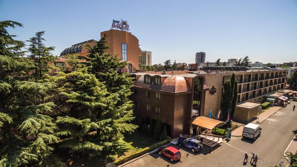 Viesnīcas ēka ar jumtu, kas rotāts ar zīmi, un apkārtējiem kokiem, skats no augšas. Gaisma ir spilgta, un redzama viesnīcas apkārtne ar automašīnām un cilvēkiem.