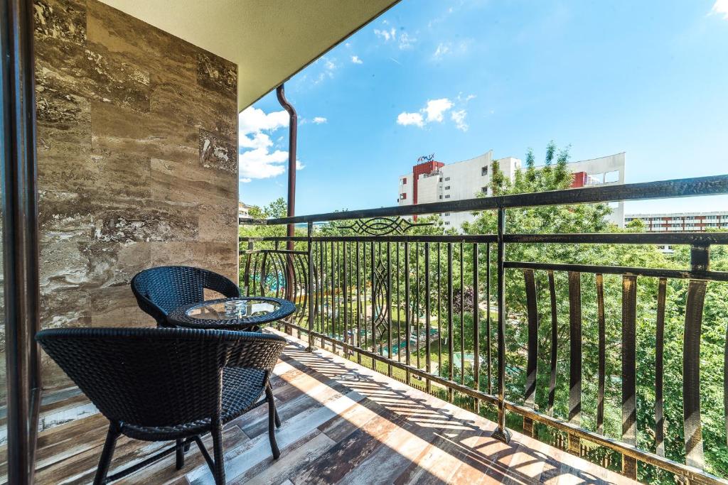 Balkons ar divām melnām krēsla un galda komplektam, kas sniedz skatu uz dārzu un apkārtējo ainavu. Saulaina diena ar zilu debesu fonu.