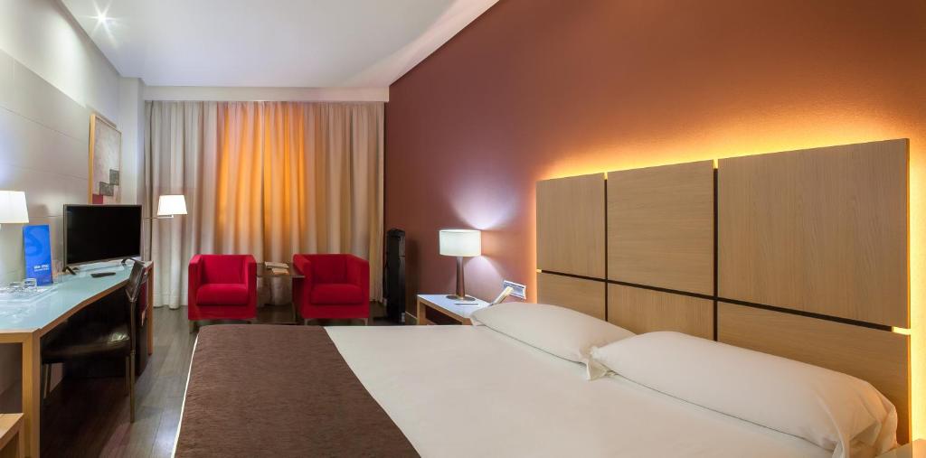 Mūsdienīgs viesnīcas numurs ar divguļamo gultu, sarkaniem krēsliem un dabiskiem apgaismojuma elementiem. Interjers apvieno komfortu un eleganci, radot patīkamu atmosfēru.