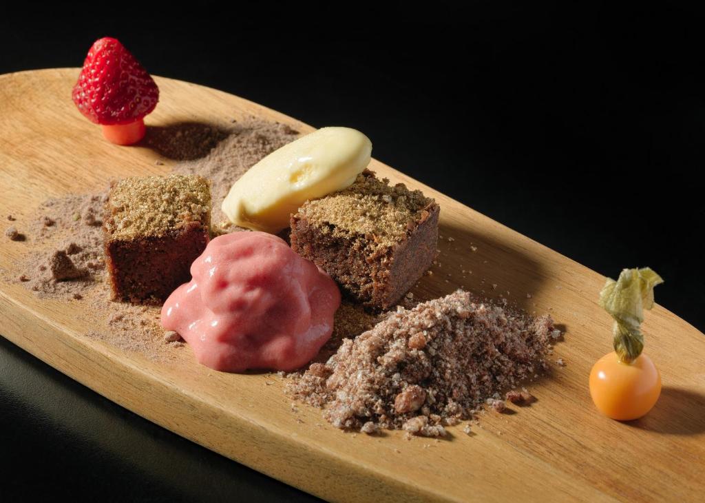 Grezns deserts ar šokolādes kūku, zemeņu saldējumu un augļu dekorācijām, pasniegts uz koka šķīvja.