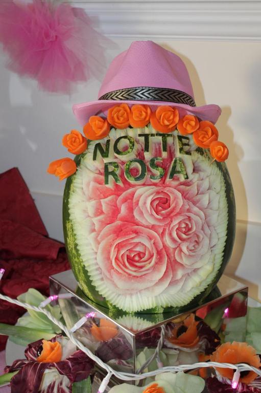 Melone ar grieztu rožu dizainu, rotāta ar oranžām ziedu dekorācijām un rozā cepuri, kas papildina svinīgo atmosfēru. Ideāli piemērota pasākumiem vai svinībām.