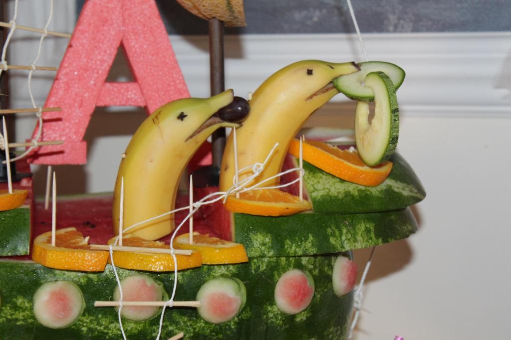 Banānu un dārzeņu figūras, kas izveidotas kā jūras dzīvnieki, uz arbūza kuģa, dekorētas ar augļiem.