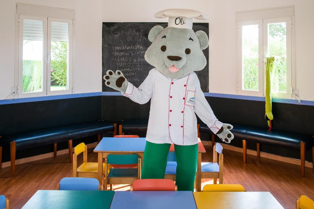 Šefpavārs lācis ar cepuri stāv virtuvē, kas aprīkota ar krāsainiem galdiem un melnu tāfeli. Aiz viņa redzami logi, caur kuriem ieplūst dabiska gaisma.