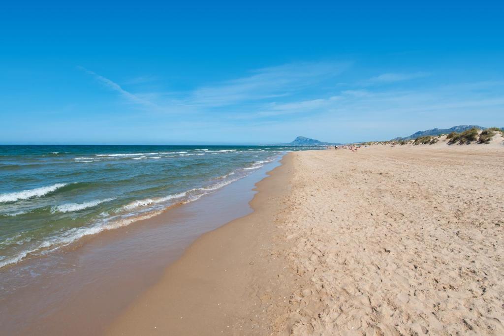 Skaists pludmales skats ar smilšu krastu un mierīgu jūru, kurā viļņi maigi saplūst ar krastu. Tālumā redzami kalni un skaidrs, saulains debesis.