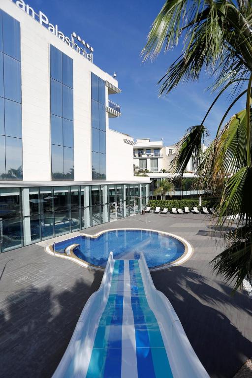 Viesnīcas āra baseins ar ūdens slidkalniņu un palmām apkārt. Modernā arhitektūra un skaidrs debesis veido relaksējošu atmosfēru.