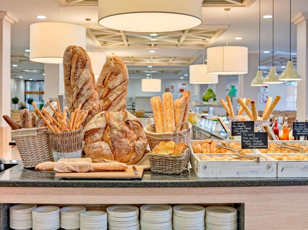 Maizes un konditorejas izstrādājumi, kas izvietoti uz galda, ar dažādām maizes šķirnēm un cepumiem, kas pasniegti grozos un kastēs. Interjers ir gaišs un mūsdienīgs, radot viesmīlīgu atmosfēru.