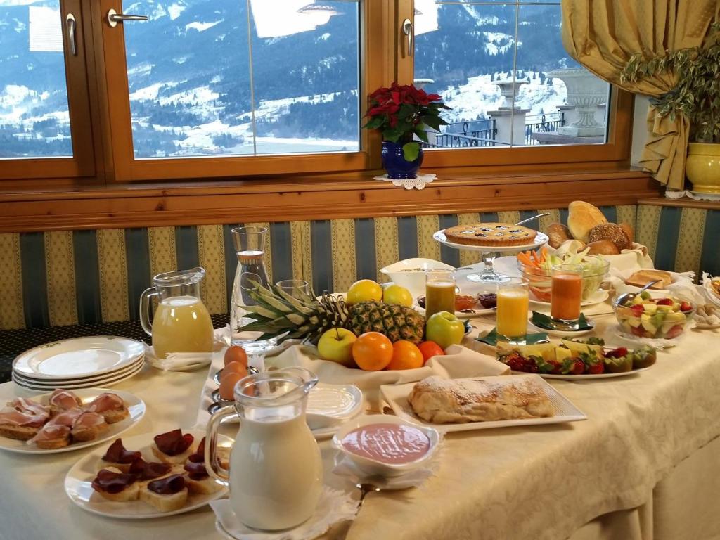 Bagātīgs brokastu galds ar augļiem, dzērieniem un dažādiem ēdieniem, kas izlikts pie loga ar kalnu skatu. Ideāla atmosfēra relaksējošām rīta maltītēm.