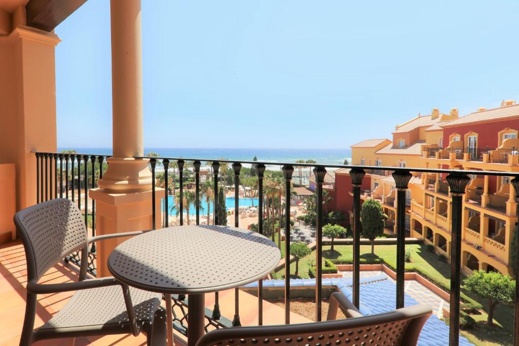 Balcony view overlooking a pool area and the ocean, featuring outdoor seating with two chairs and a table, surrounded by lush greenery and colorful buildings.