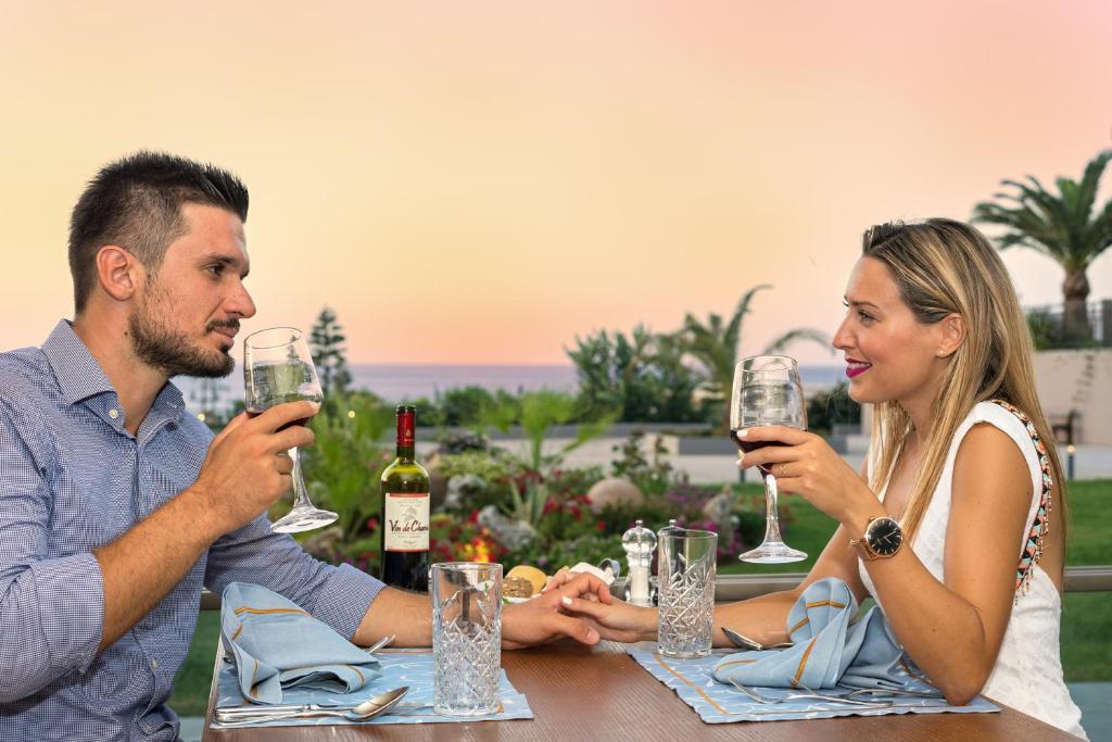 Romantiska vakariņas pāri pie galda ar dzērieniem, skats uz jūru un saulrietu. Galda klājums ar zilu galdautu un vīna pudeli.