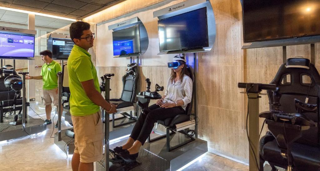 Virtuālās realitātes spēļu zona ar lietotājiem, kas izmanto VR austiņas un specializētas sēdvietas, lai izbaudītu spēles. Vides moderns dizains ar ekrāniem un tehnoloģijām.