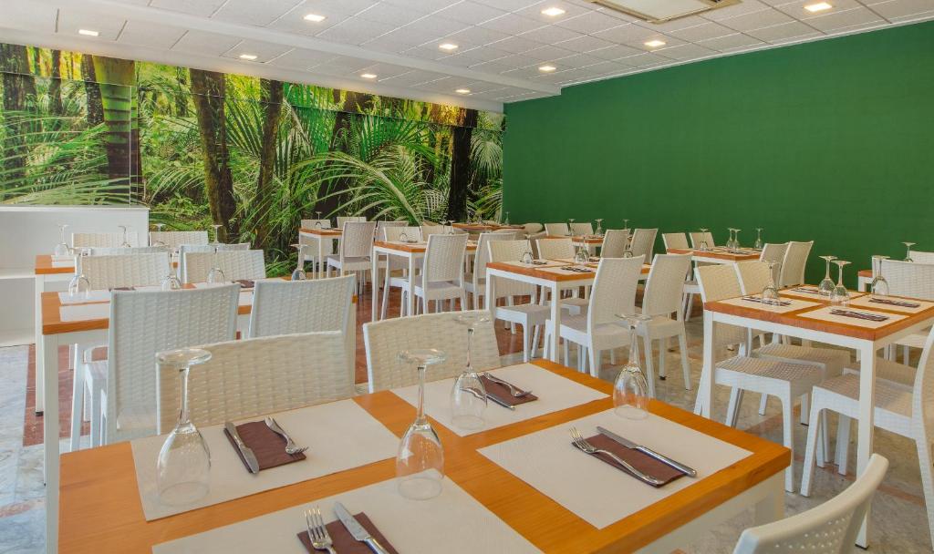 Mūsdienīgs restorāns ar elegantām baltām mēbelēm un zaļiem akcentiem, kas radīts, lai nodrošinātu patīkamu ēšanas pieredzi. Fons ar tropu augu attēlu piešķir telpai dzīvīgumu.