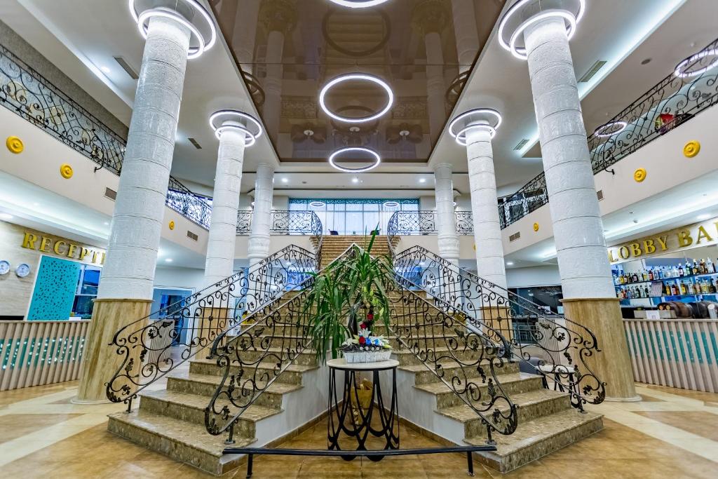 Elegants viesnīcas vestibils ar plašu kāpņu telpu, dekoratīvām margām un mūsdienīgiem gaismas elementiem. Redzama reģistratūras zona un bārs.