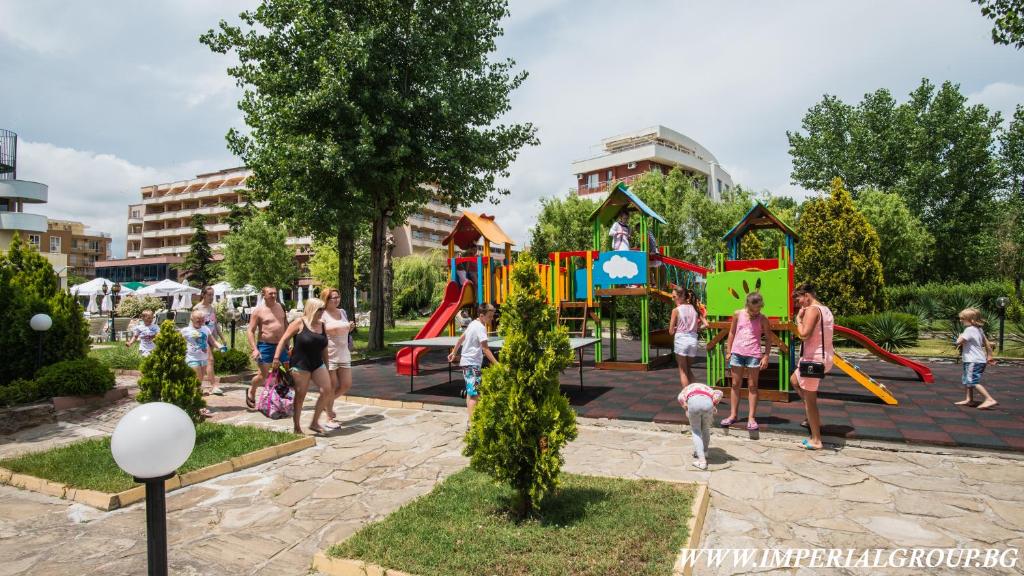 Bērnu spēļu laukums ar krāsainām struktūrām, kurā bērni rotaļājas un pieaugušie pastaigājas pa zaļojošo teritoriju. Apkārt ir koki un ziedi, radot patīkamu atmosfēru.