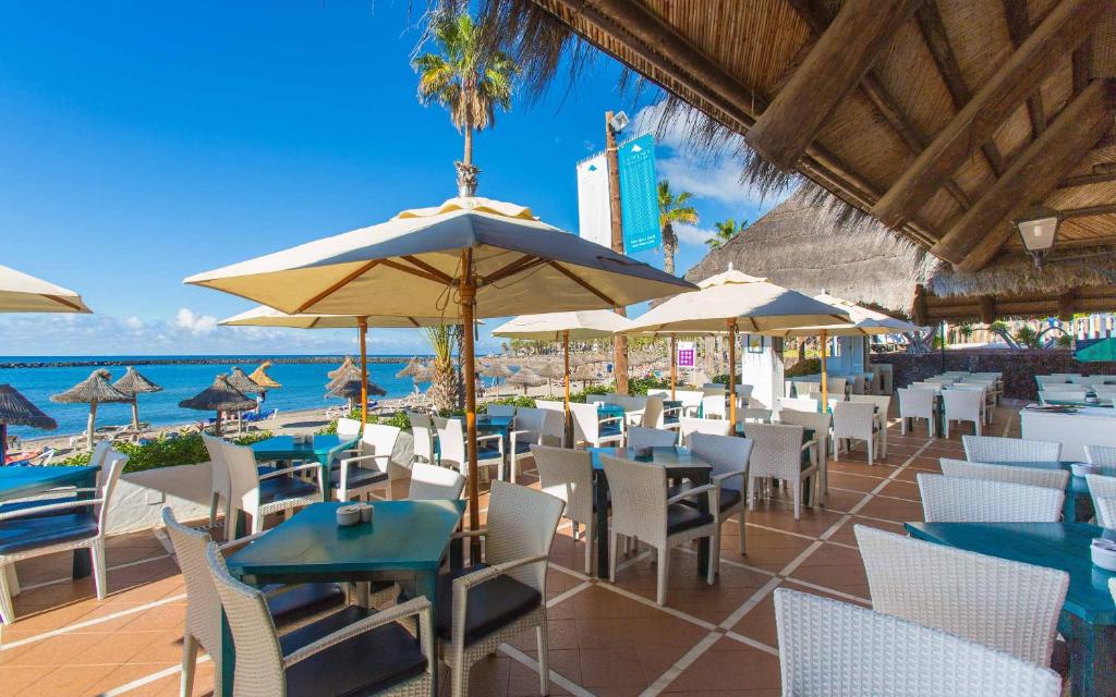 Atklāta terase pie pludmales ar baltiem galdiem un krēsliem, saulessargi un zila jūra fona. Ideāla vieta relaksācijai un ēdienreizei brīvā dabā.